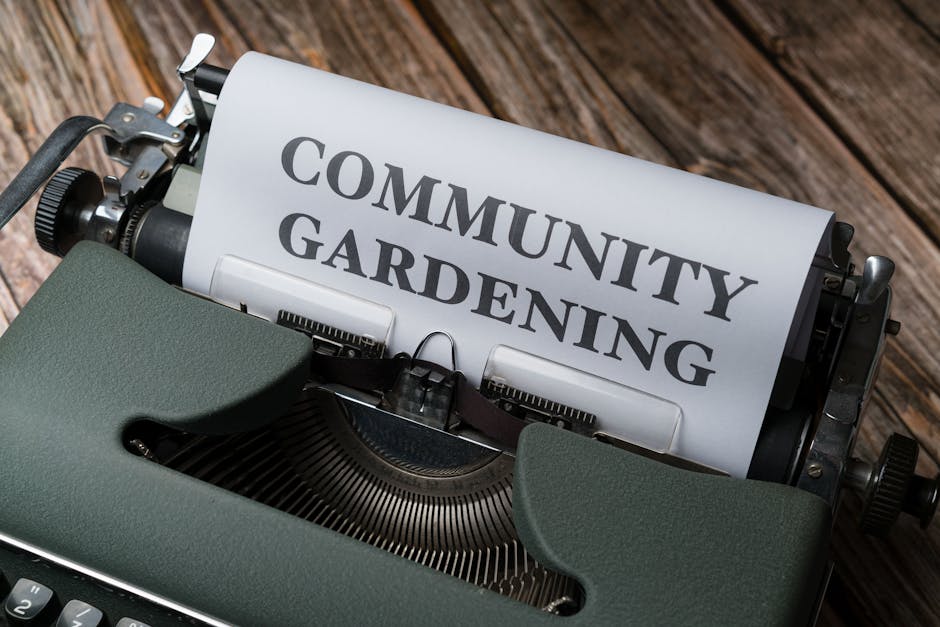 A vintage green typewriter resting on a wooden surface, with a sheet of paper inserted that displays the words 'Community Gardening' in bold, dark grey letters. The background shows weathered, texture