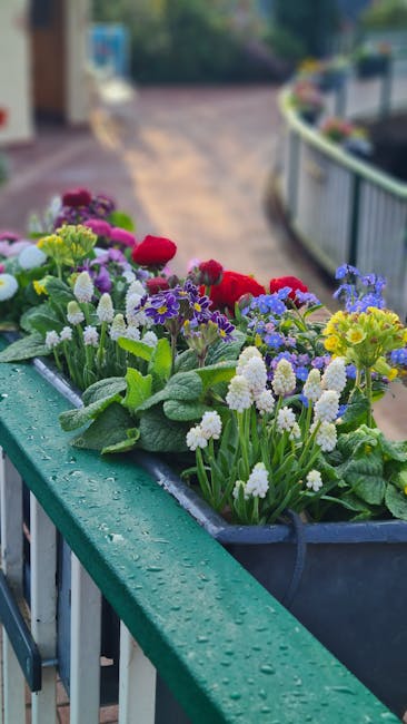 A close-up view of a balcony garden planter box filled with a variety of colorful flowering plants, including red, purple, and white blooms, with lush green leaves. The container appears to be made of