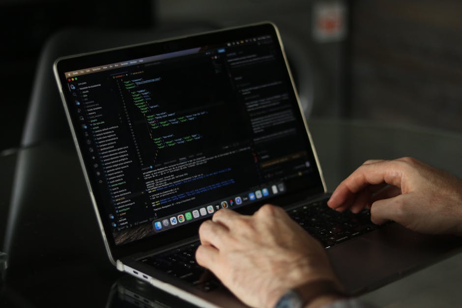 A close-up view of a person’s hands typing on a MacBook laptop situated on a dark surface, with the screen displaying lines of computer programming code in various colors on a dark background. In th